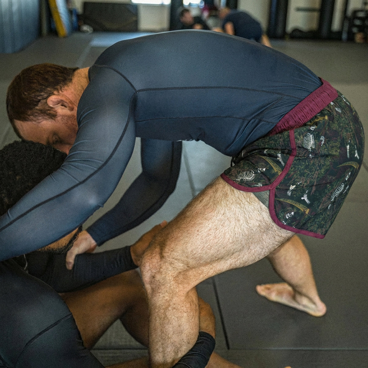 Man wearing ZF Fight Shorts with a camouflage pattern and maroon trim practicing grappling on a mat with a training partner in a gym setting.