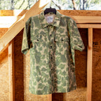Camo-patterned short sleeve CPO Shirt hanging on a black hanger against a wooden framed wall with natural light highlighting the fabric texture.