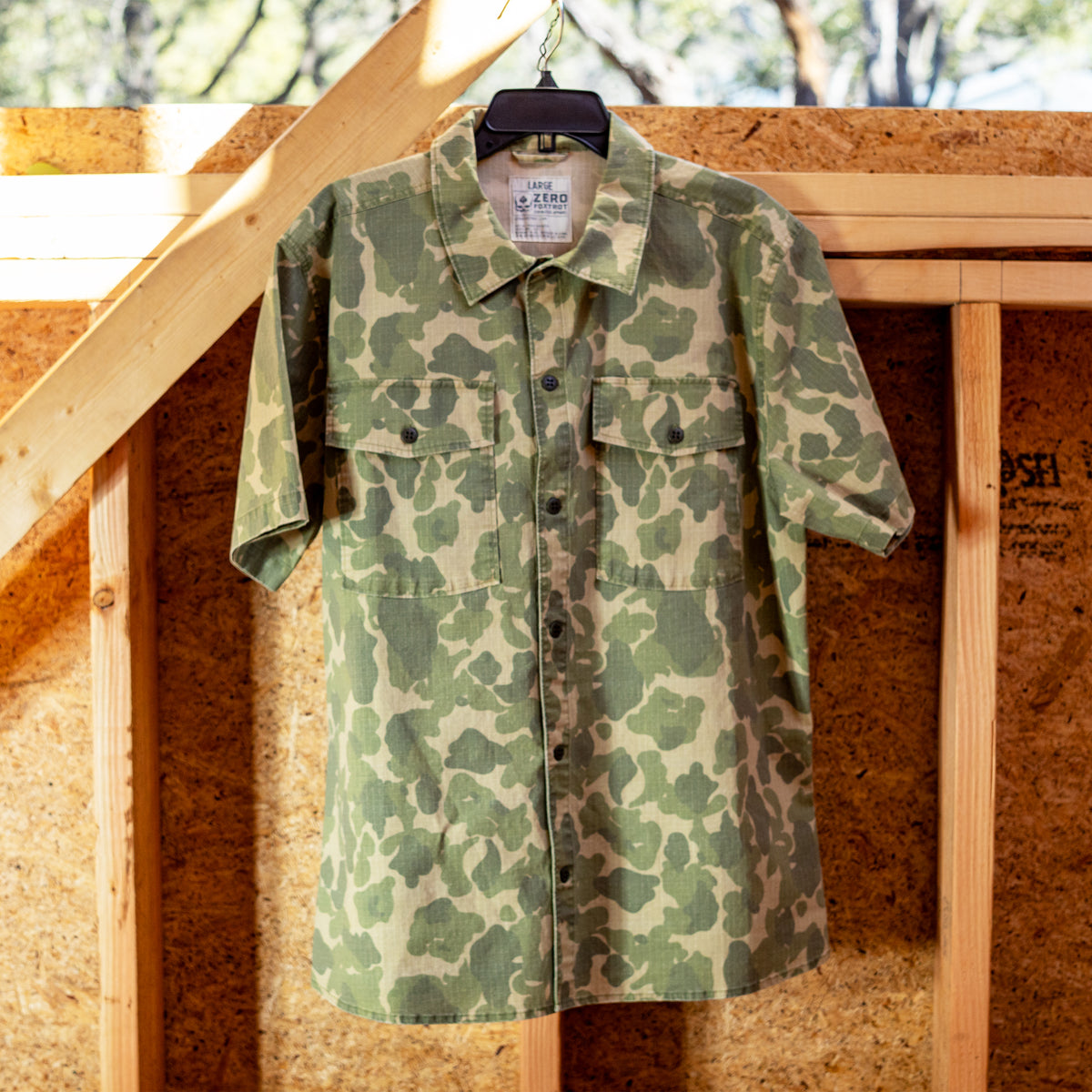 Camo-patterned short sleeve CPO Shirt hanging on a black hanger against a wooden framed wall with natural light highlighting the fabric texture.