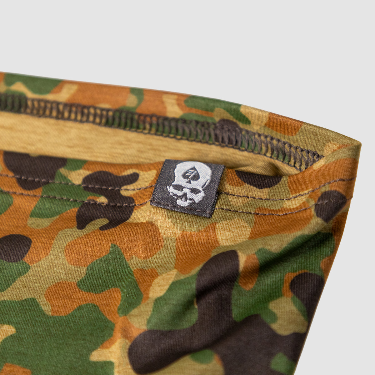 Close-up of a camo-patterned Performance Sun Gaiter made from lightweight fabric featuring durable stitching and a small black label with a white skull and spade logo.