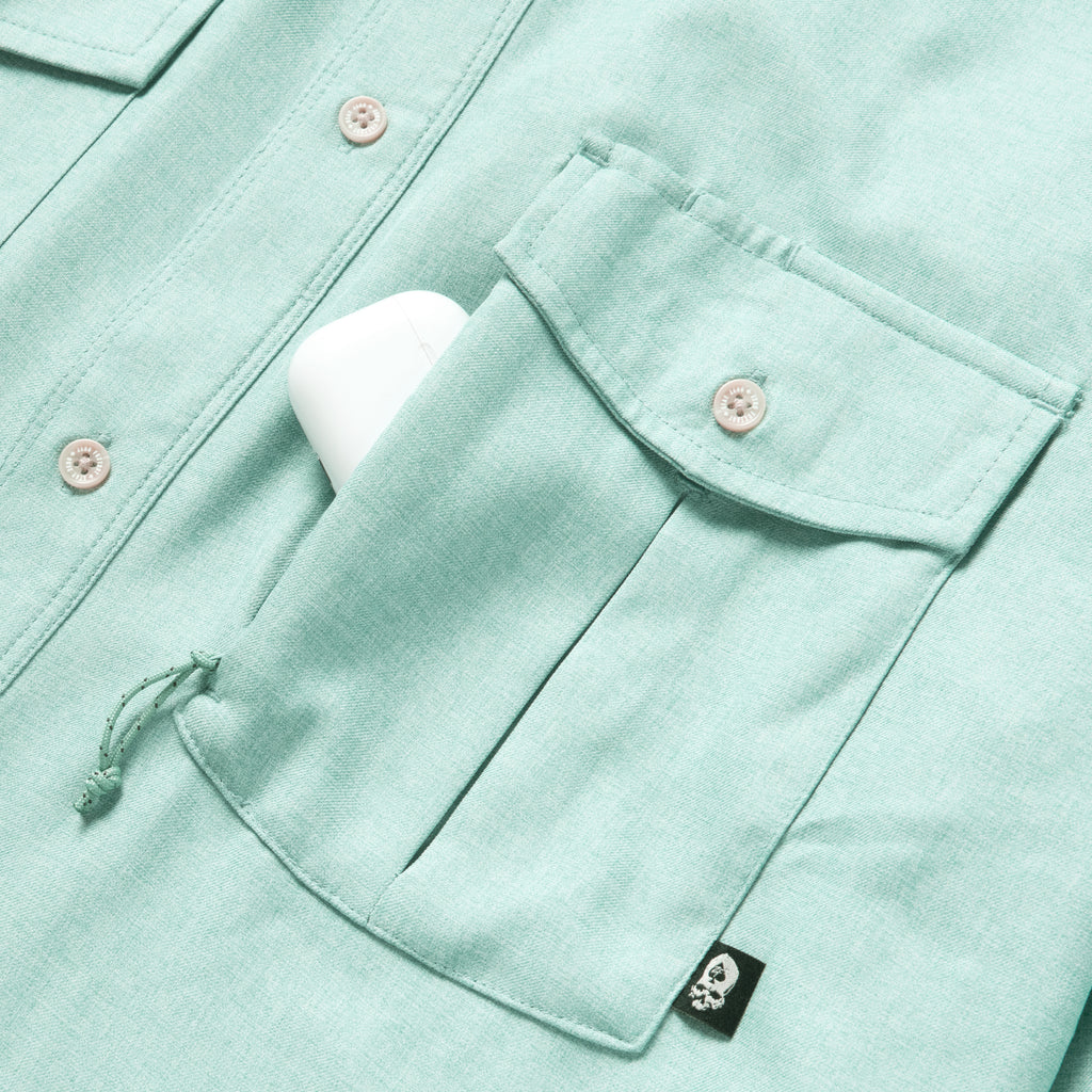 Close-up of a light aqua F'n Soft Daily Shirt pocket with a button closure holding a white item, showing detailed stitching and a small black skull tag.
