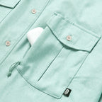 Close-up of a light aqua F'n Soft Daily Shirt pocket with a button closure holding a white item, showing detailed stitching and a small black skull tag.