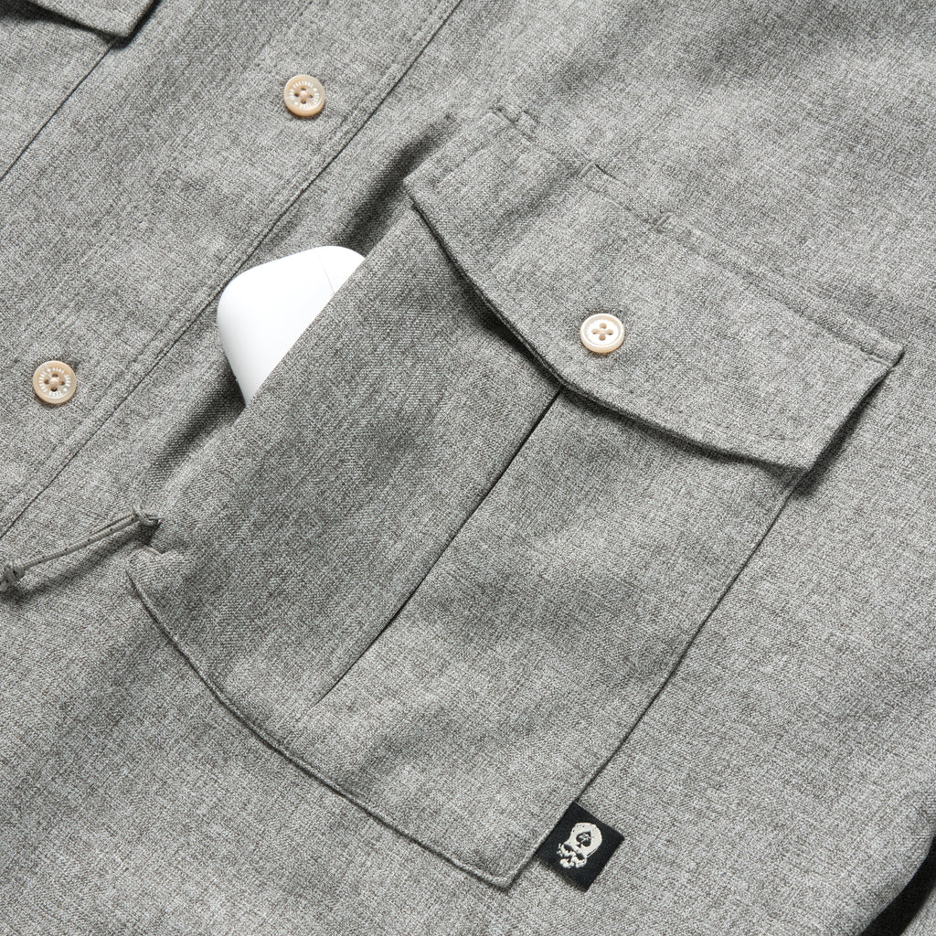Close-up of a charcoal gray textured fabric pocket with button closure and a small black skull tag detail on the F'n Soft Daily Shirt, showing part of the button-up front.