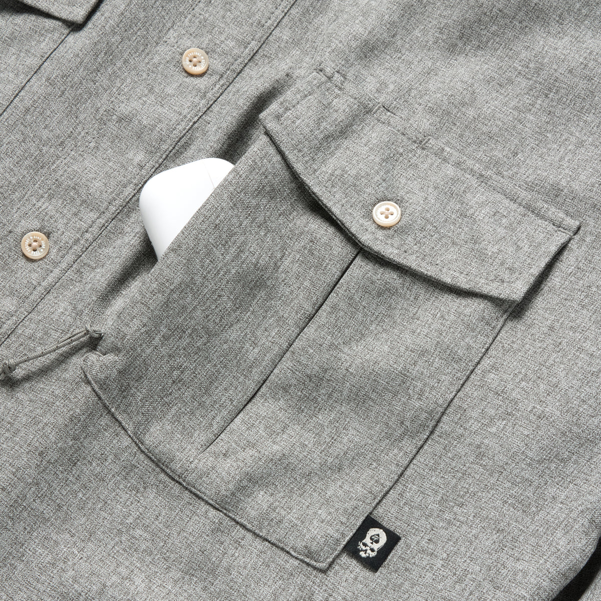 Close-up of a charcoal gray textured fabric pocket with button closure and a small black skull tag detail on the F'n Soft Daily Shirt, showing part of the button-up front.