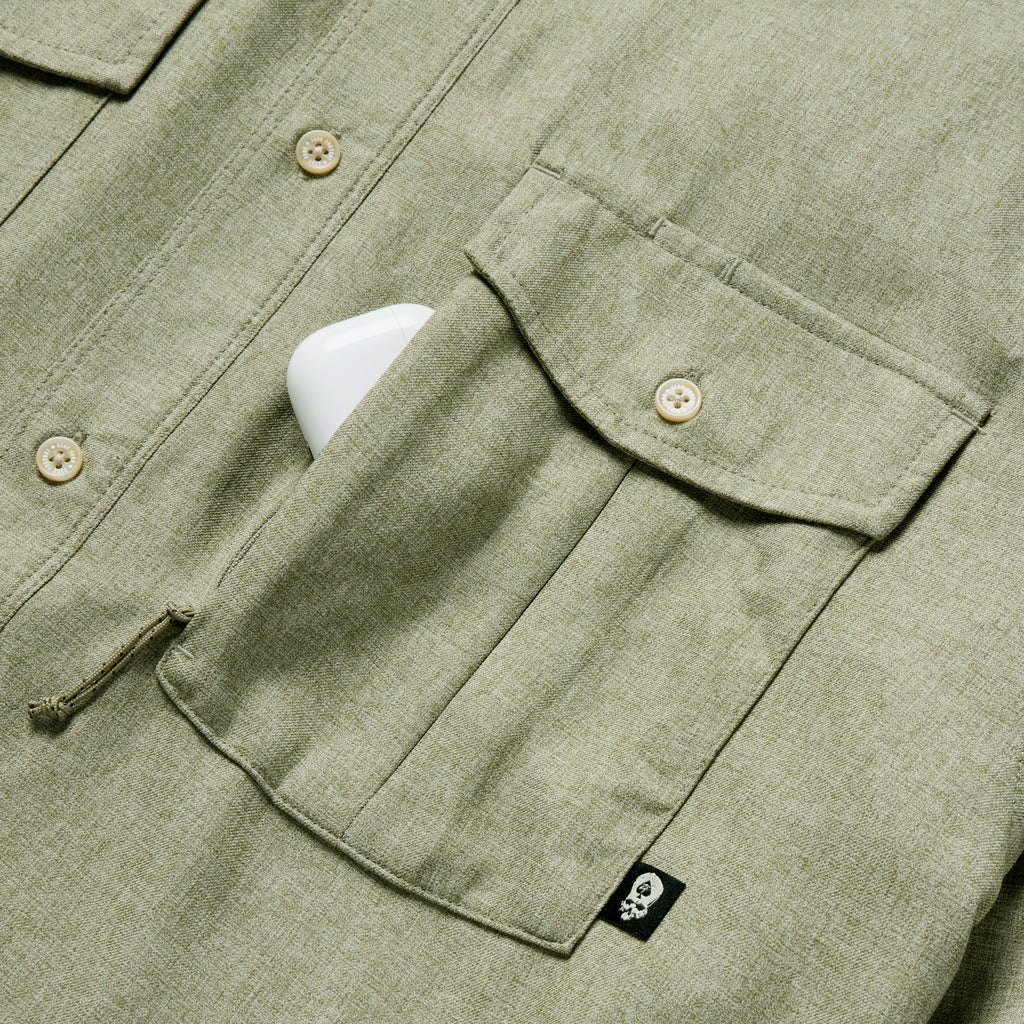 Close-up of a sage green F'n Soft Daily Shirt showing button details and a chest pocket with a small black tag, partially holding a white phone.