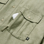 Close-up of a sage green F'n Soft Daily Shirt showing button details and a chest pocket with a small black tag, partially holding a white phone.