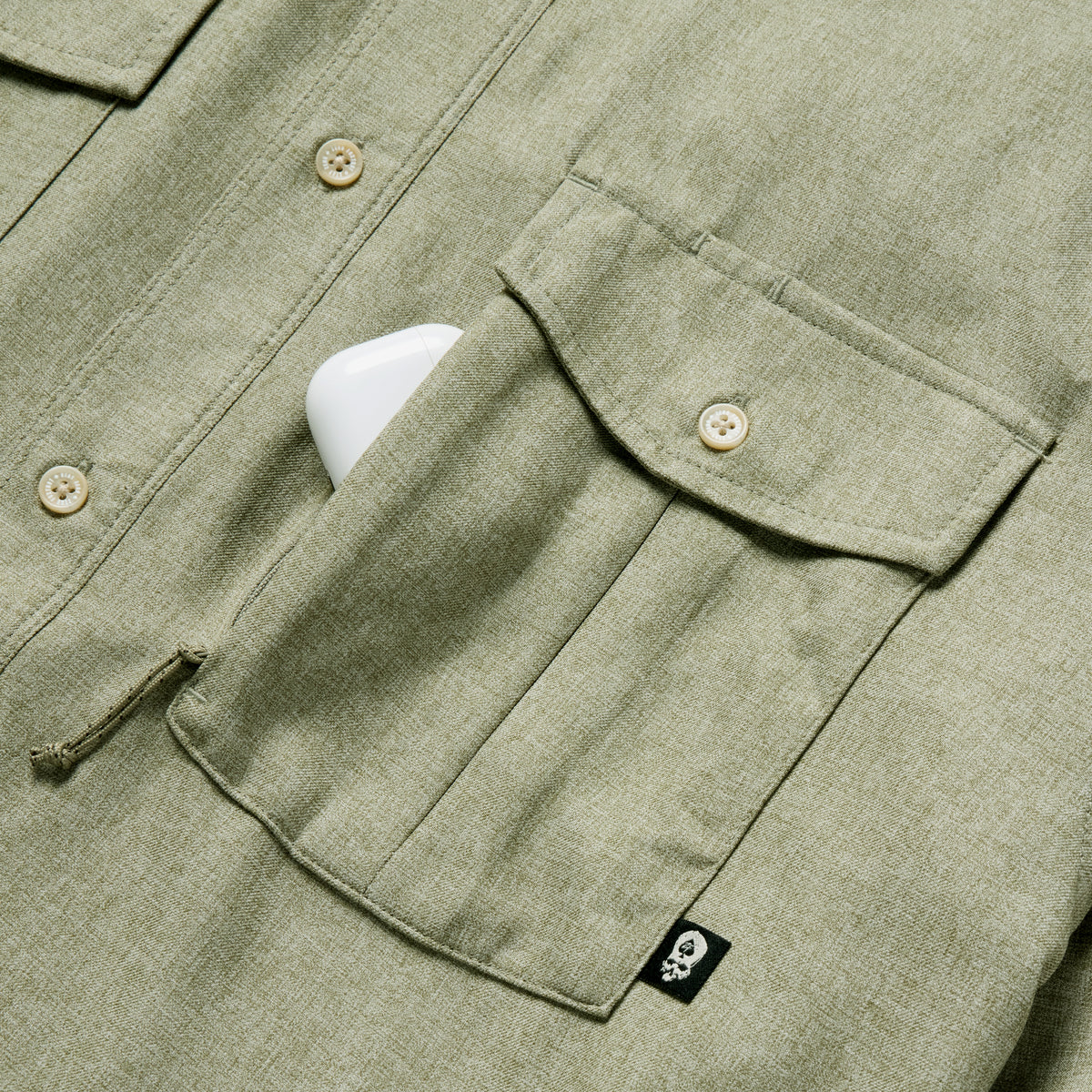 Close-up of a sage green F'n Soft Daily Shirt showing button details and a chest pocket with a small black tag, partially holding a white phone.