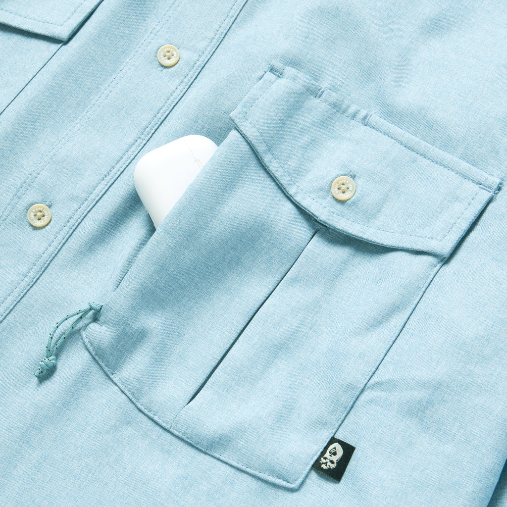 Close-up of a light blue F'n Soft Daily Shirt pocket with a button flap and a small black skull tag, showing a white object partially tucked inside the pocket.