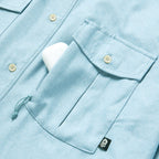 Close-up of a light blue F'n Soft Daily Shirt pocket with a button flap and a small black skull tag, showing a white object partially tucked inside the pocket.