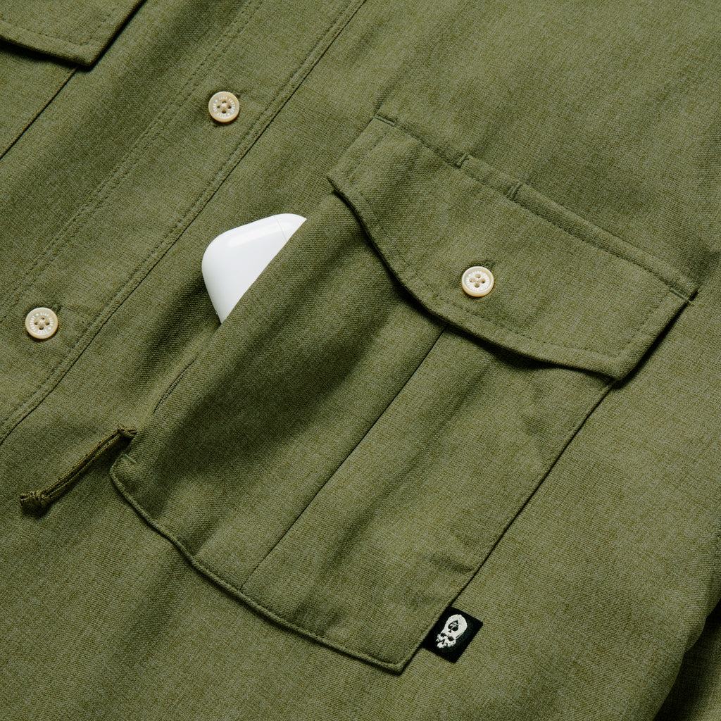 Close-up of an olive green F'n Soft Daily Shirt pocket with button closure and a small black tag featuring a white skull, partially holding a white object.