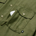 Close-up of an olive green F'n Soft Daily Shirt pocket with button closure and a small black tag featuring a white skull, partially holding a white object.
