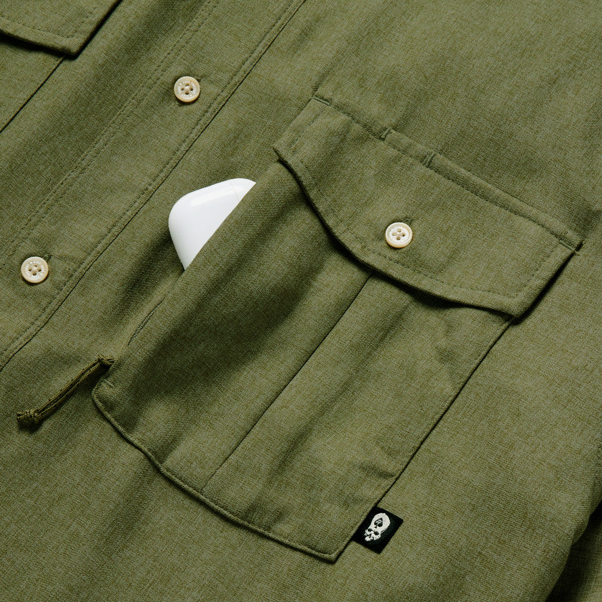 Close-up of an olive green F'n Soft Daily Shirt pocket with button closure and a small black tag featuring a white skull, partially holding a white object.