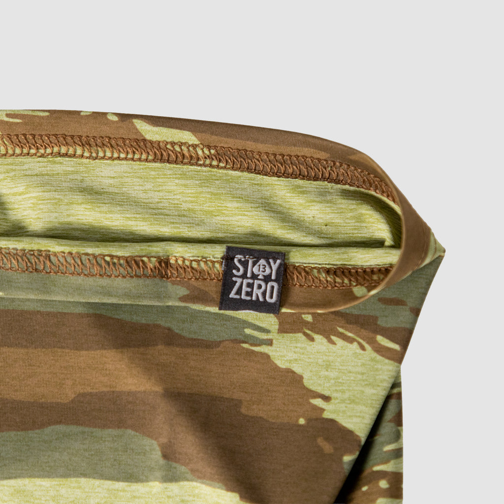 Close-up of a camouflage patterned fabric with a stitched edge and a small black label, showing the Performance Sun Gaiter designed for sun protection and outdoor use.