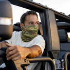 Man wearing a camouflage Performance Sun Gaiter covering his face and neck while sitting in an open-top vehicle during daylight.
