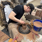 Person shaping clay on a pottery wheel in a workshop, crafting a Zero Dark Mug with tools and a bucket of wet clay nearby.