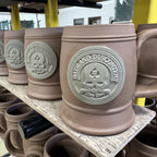 Row of unglazed ceramic mugs on a wooden shelf, each featuring a raised emblem with "RAVE AND DISCOVERY" text, showcasing the design on the Zero Dark Mug before firing.