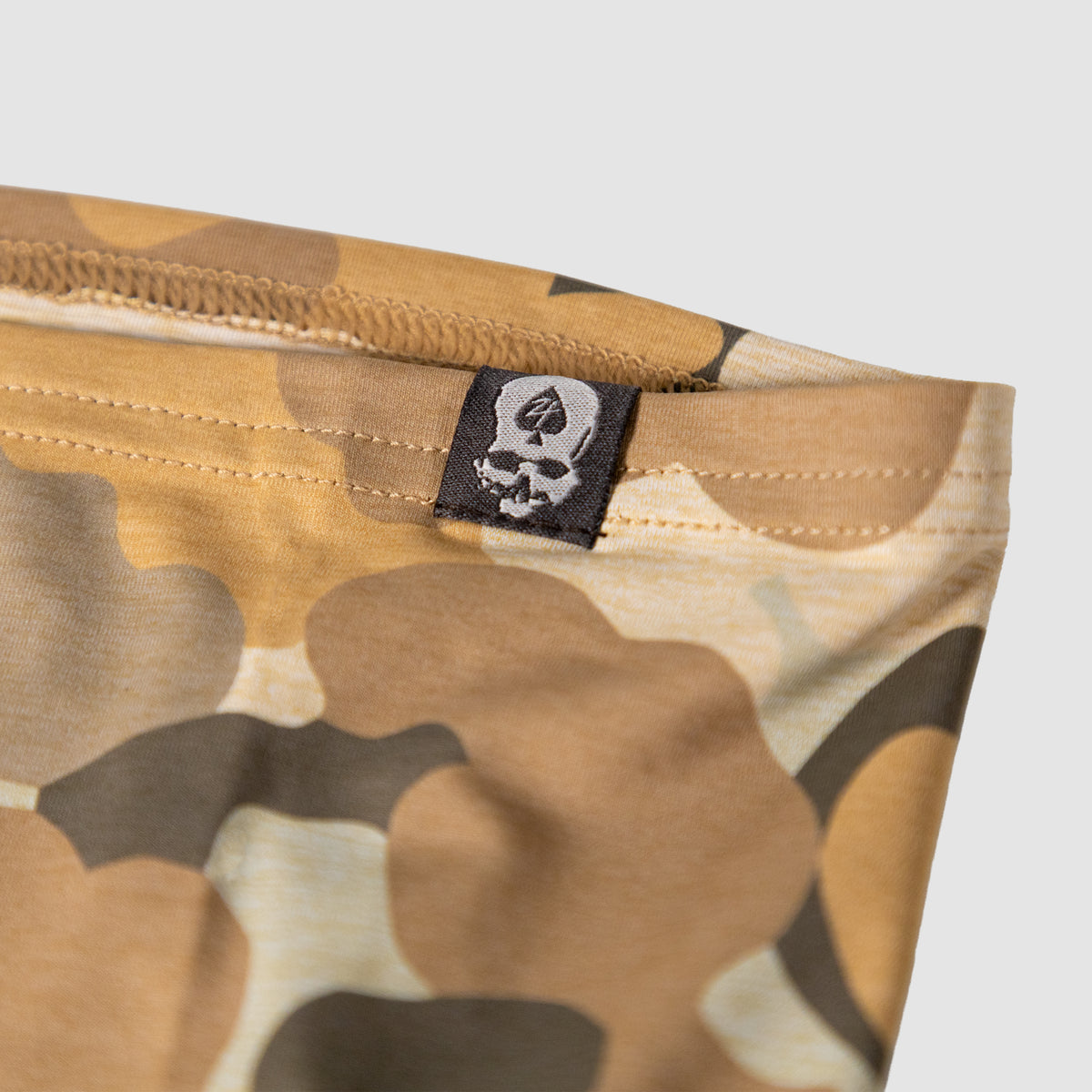 Close-up of a tan and brown camouflage fabric with detailed stitching and a small black tag featuring a skull logo on the Performance Sun Gaiter.