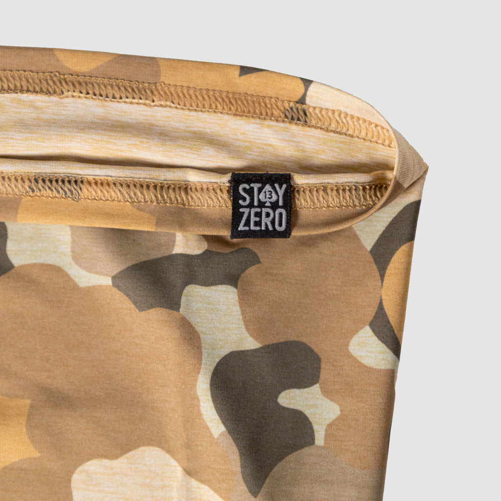 Close-up of a tan and brown camouflage-patterned Performance Sun Gaiter with a visible black tag featuring white text, highlighting the fabric texture and stitching details.