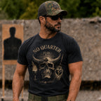 Man wearing sunglasses and a camo cap with a skull and horn graphic design on a black No Quarter Ghost Tee t-shirt, standing outdoors in a shooting range.