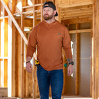Man wearing a rust-colored Performance Sun Hoodie with a front pocket, jeans, and a backward cap, walking inside a wooden framed construction site.
