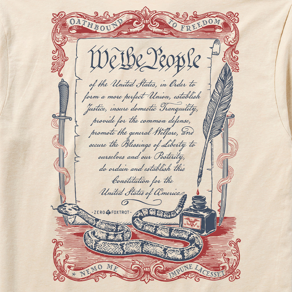 Oathbound Tee featuring a vintage-style print of the U.S. Constitution's preamble with decorative red and blue scrollwork, a coiled rattlesnake, ink bottle, quill pen, and sword on a light beige fabric.