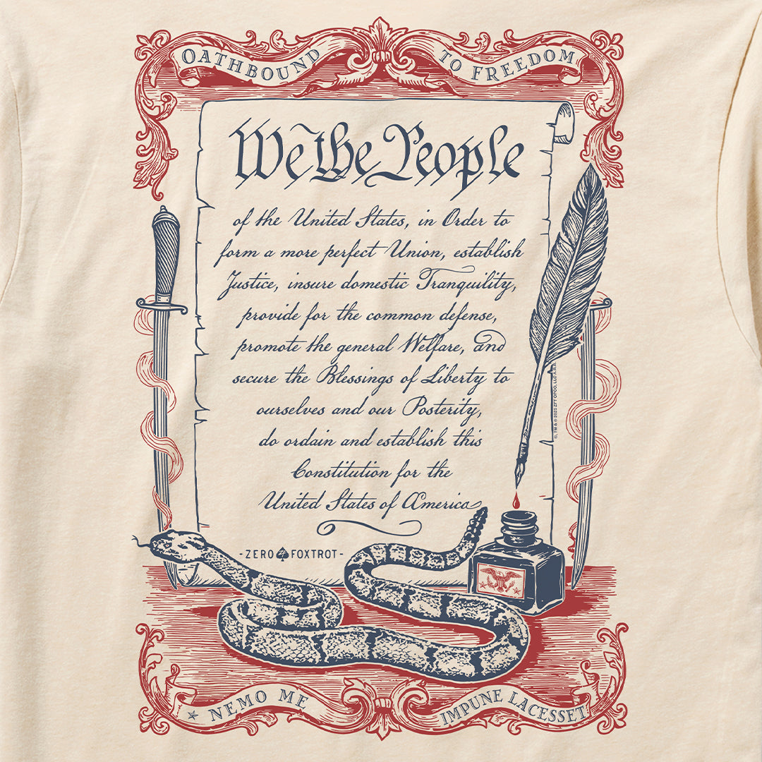 Oathbound Tee featuring a vintage-style print of the U.S. Constitution's preamble with decorative red and blue scrollwork, a coiled rattlesnake, ink bottle, quill pen, and sword on a light beige fabric.