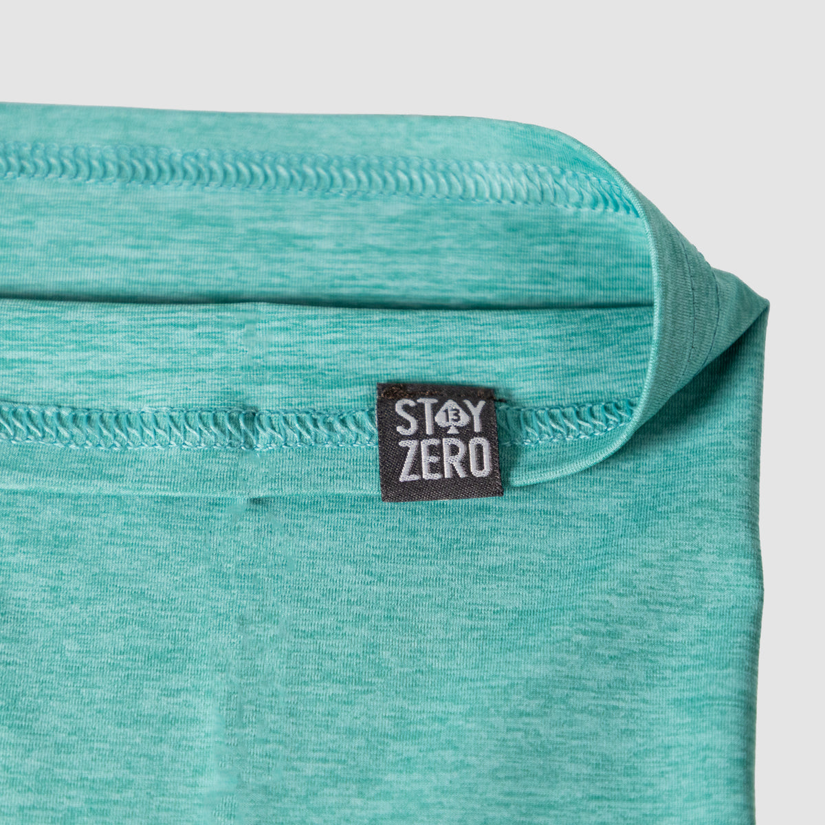 Close-up view of a turquoise fabric Performance Sun Gaiter with a visible black tag labeled Stay Zero, highlighting the material texture and stitching details.