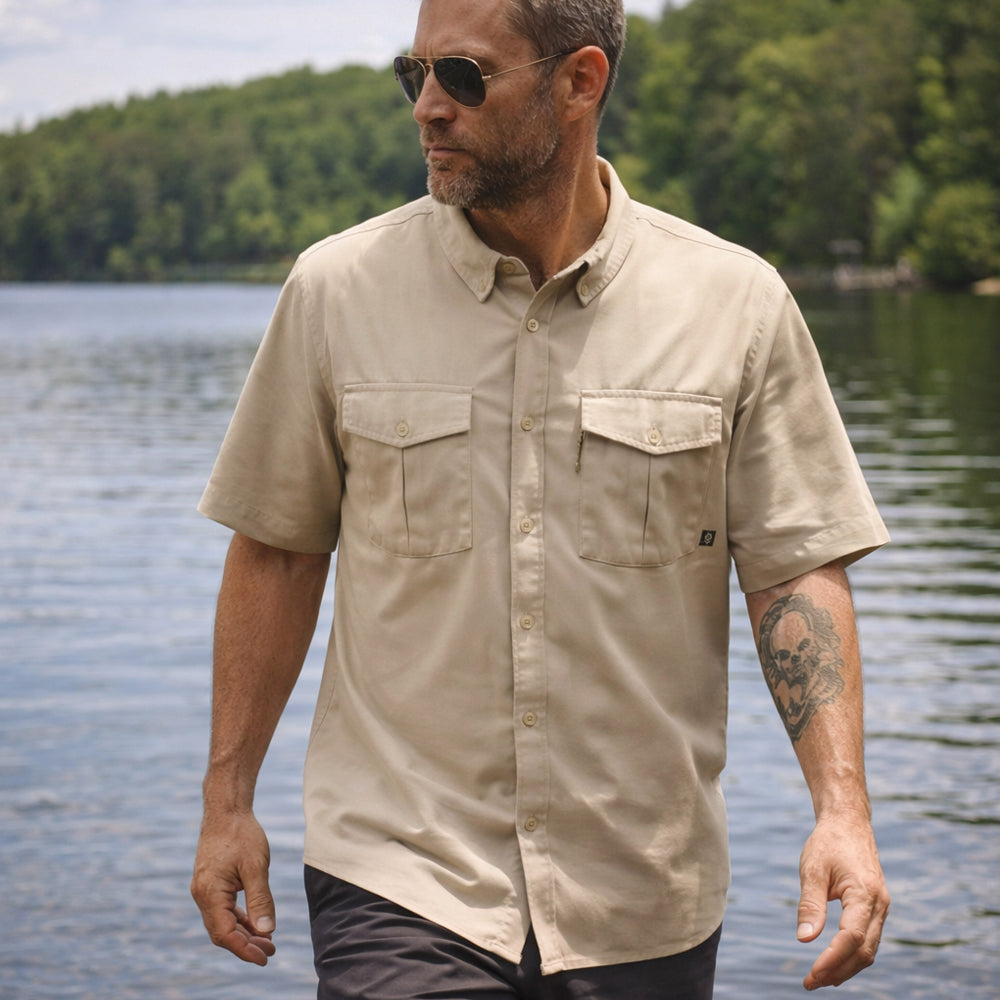 Man wearing a tan short sleeve F'n Soft Shirt with buttoned chest pockets and sunglasses, standing outdoors near a body of water with trees in the background.