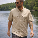 Man wearing a tan short sleeve F'n Soft Shirt with buttoned chest pockets and sunglasses, standing outdoors near a body of water with trees in the background.