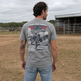 Man wearing a gray Feel the Need Tee featuring a vintage aviation graphic with a pilot and fighter jets, standing outdoors in a rural setting near a fenced area.