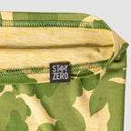 Close-up of a green and yellow camouflage Performance Sun Gaiter showing the fabric texture and a black sewn-in tag with the text "STAY ZERO.