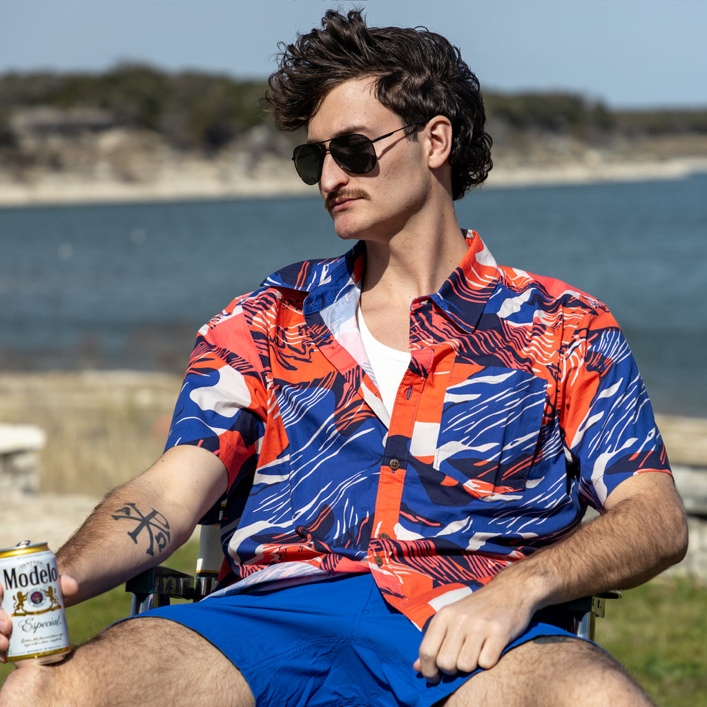 Surf Shirts (Last Chance) featuring a man in a red and blue patterned short sleeve button shirt, seated outdoors by the water in sunglasses and blue shorts