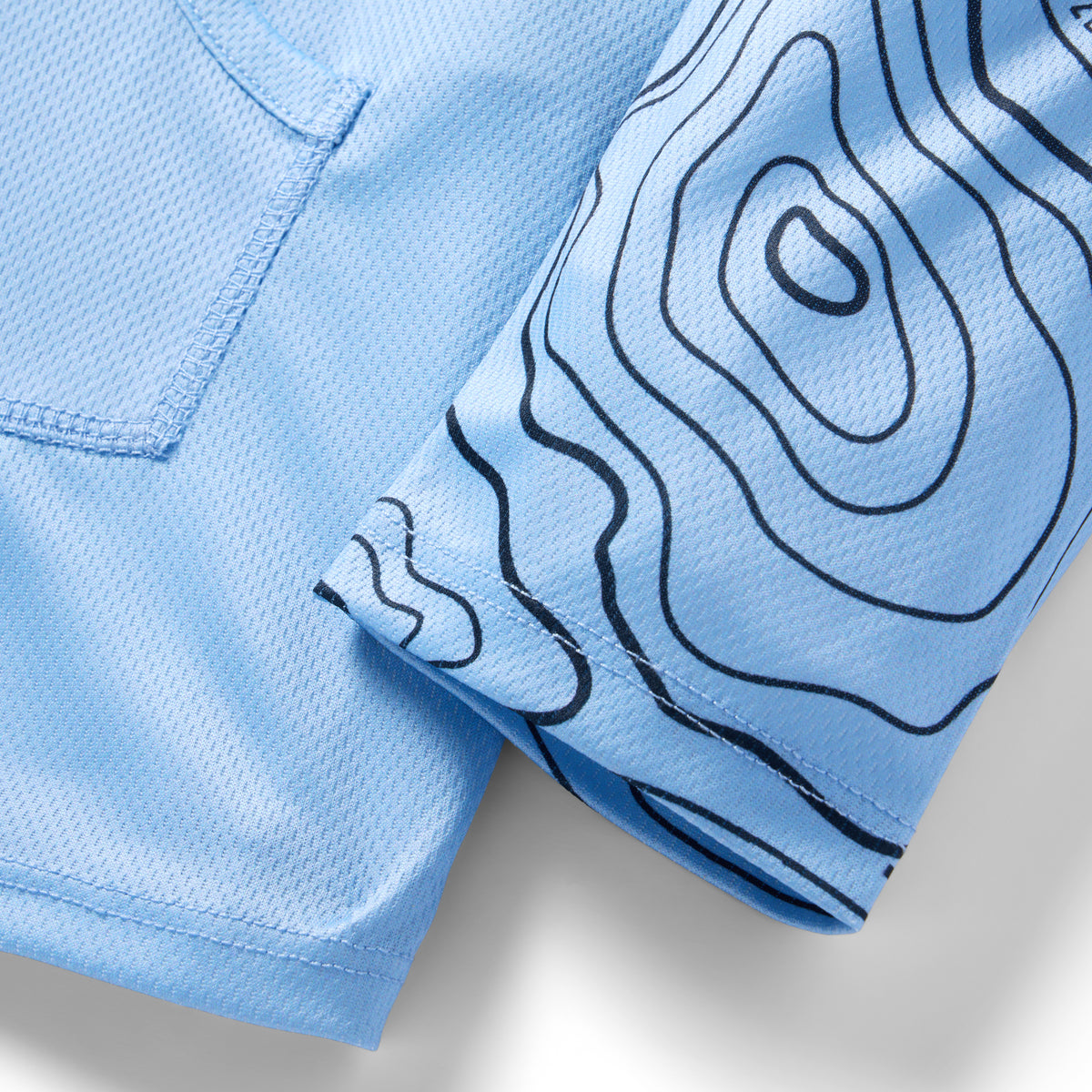 Close-up of light blue fabric with topographic line patterns on the sleeve and mesh texture on the body of the Crew Rash Guard (Last Season) shirt.