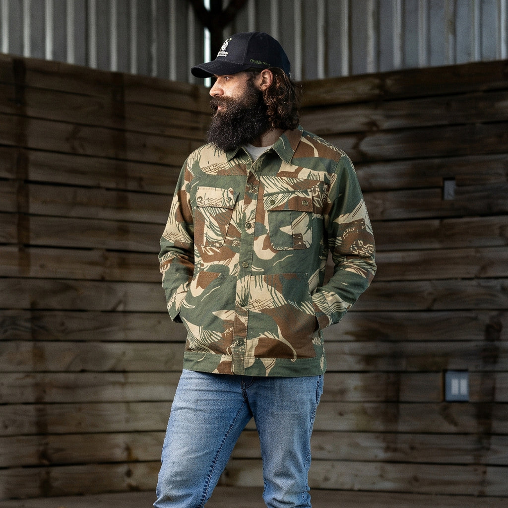Man wearing a camouflage pattern Chore Jacket with front pockets, paired with light blue jeans and a black baseball cap, standing in a rustic wooden interior.