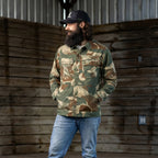 Man wearing a camouflage pattern Chore Jacket with front pockets, paired with light blue jeans and a black baseball cap, standing in a rustic wooden interior.