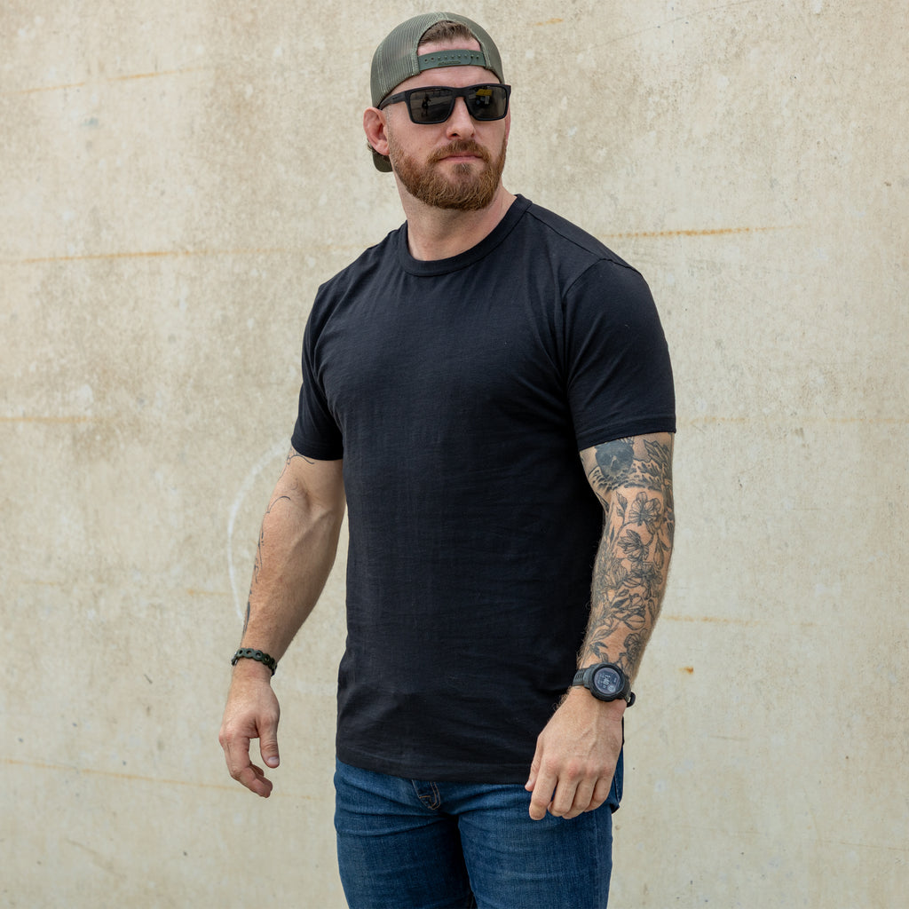 Man wearing a black Cotton Skivvy Tee with sunglasses, a backward cap, blue jeans, and visible arm tattoos standing against a plain beige wall.