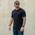 Man wearing a black Cotton Skivvy Tee with sunglasses, a backward cap, blue jeans, and visible arm tattoos standing against a plain beige wall.