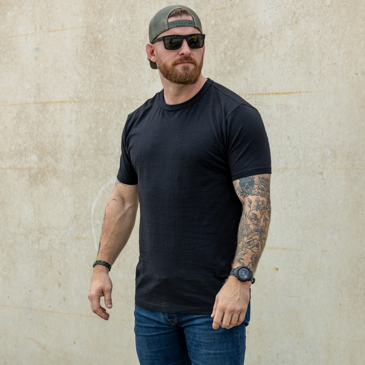 Man wearing a black Cotton Skivvy Tee with sunglasses, a backward cap, blue jeans, and visible arm tattoos standing against a plain beige wall.