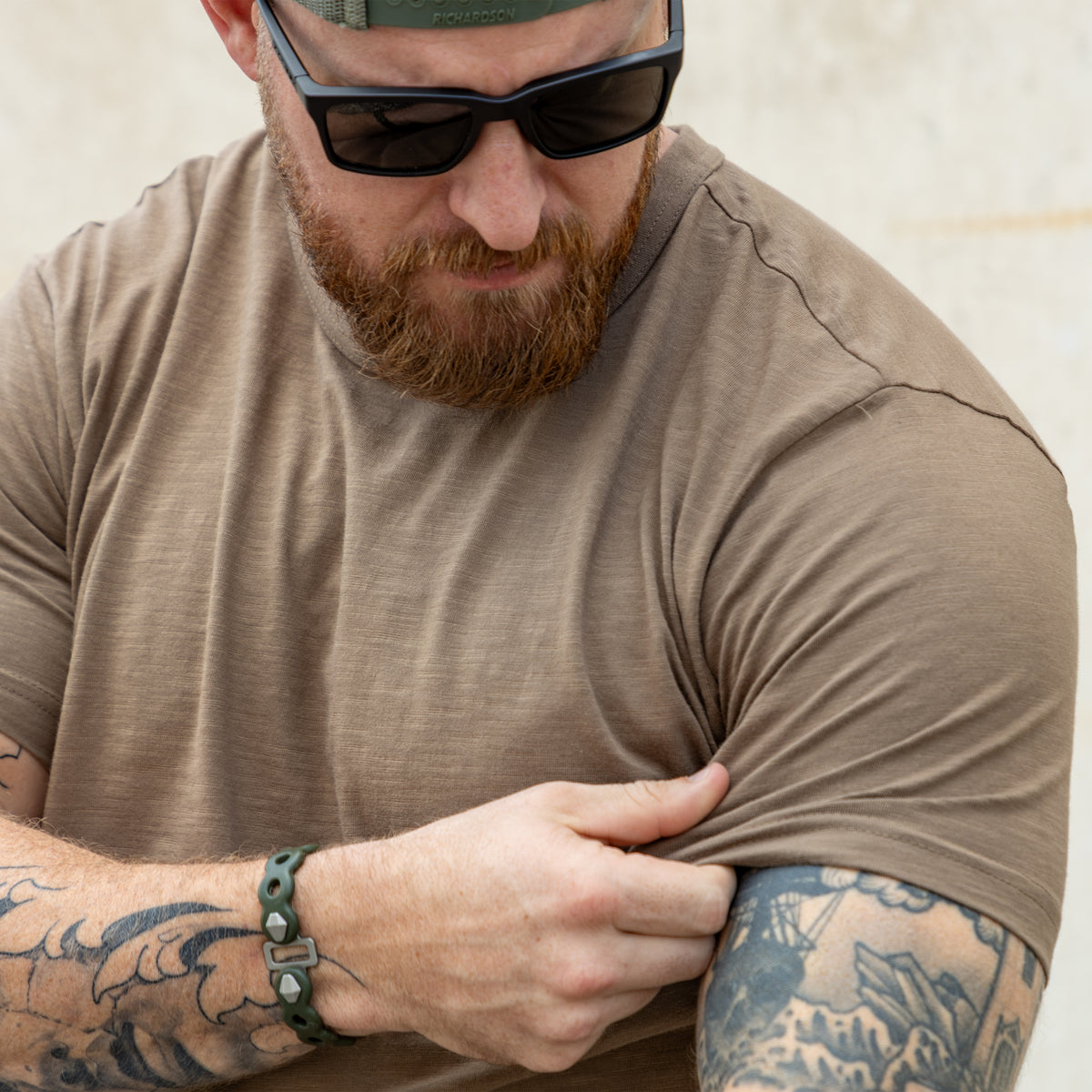 Man wearing a light brown Cotton Skivvy Tee, pulling at the sleeve to show the fabric, with sunglasses and visible tattoos on his arms.
