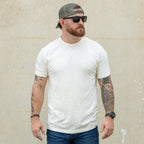 Man wearing a fitted cream-colored Cotton Skivvy Tee with short sleeves, paired with blue jeans, sunglasses, a green cap worn backwards, and visible arm tattoos.