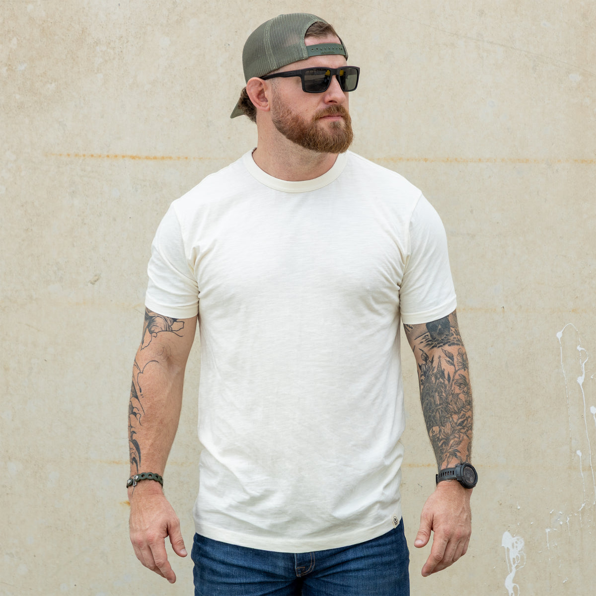 Man wearing a fitted cream-colored Cotton Skivvy Tee with short sleeves, paired with blue jeans, sunglasses, a green cap worn backwards, and visible arm tattoos.