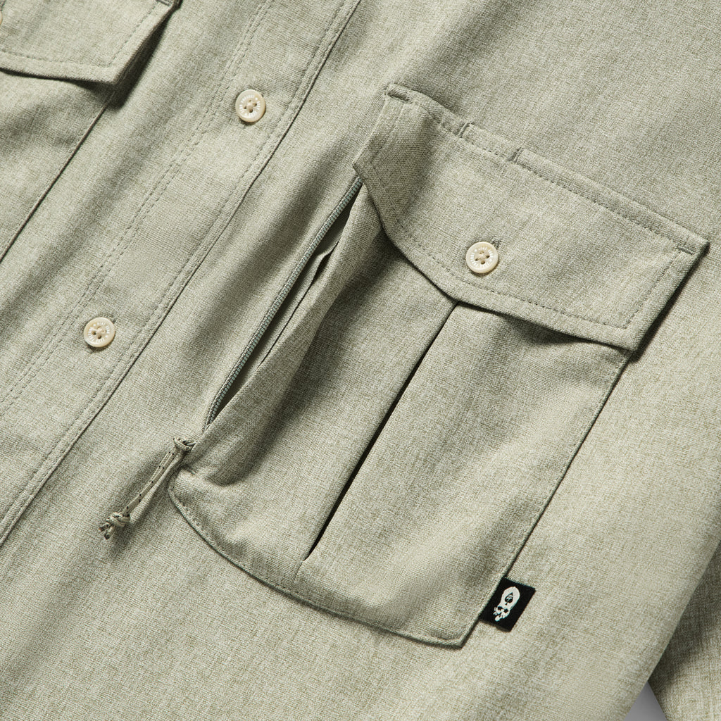 Close-up of a sage green Short Sleeve F'n Soft Shirt showing a buttoned flap pocket with a zipper underneath and a small black tag with a skull logo.