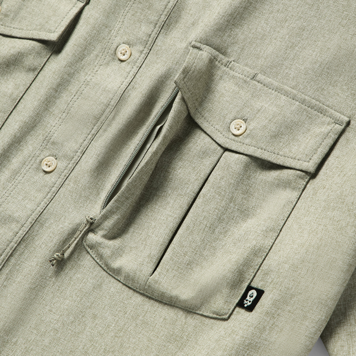 Close-up of a sage green Short Sleeve F'n Soft Shirt showing a buttoned flap pocket with a zipper underneath and a small black tag with a skull logo.