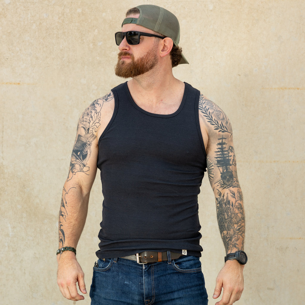 Man wearing a black Skivvy Tank, blue jeans, sunglasses, and a backwards cap, standing against a light-colored wall with tattooed arms visible.