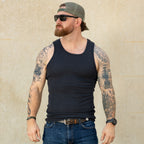 Man wearing a black Skivvy Tank, blue jeans, sunglasses, and a backwards cap, standing against a light-colored wall with tattooed arms visible.