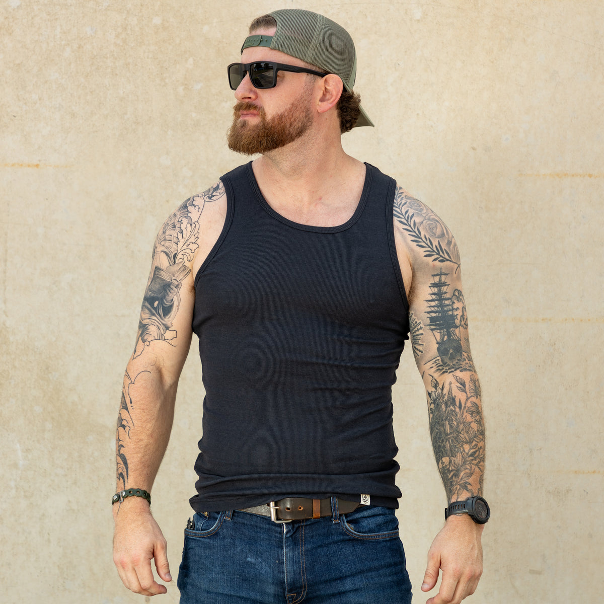 Man wearing a black Skivvy Tank, blue jeans, sunglasses, and a backwards cap, standing against a light-colored wall with tattooed arms visible.
