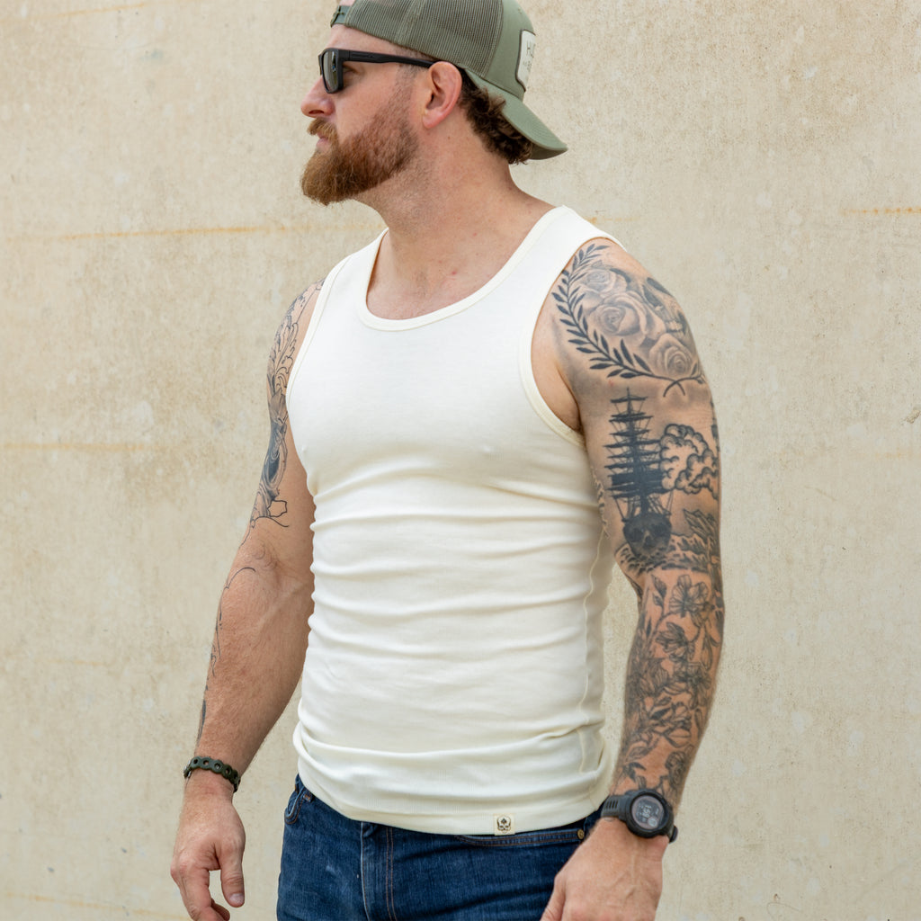 Man wearing a fitted off-white Skivvy Tank with a round neckline, paired with jeans, a green backward cap, sunglasses, and showing intricate arm tattoos.