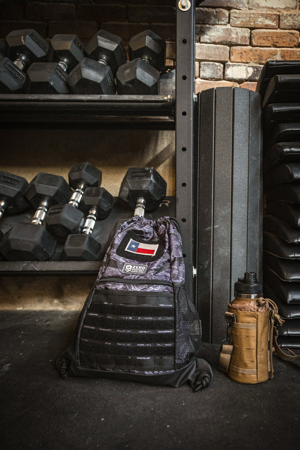 TAC Drawstring Pack in a gym setting, placed on the floor near dumbbells and exercise mats, featuring a camouflage design with a patch detail on the front.