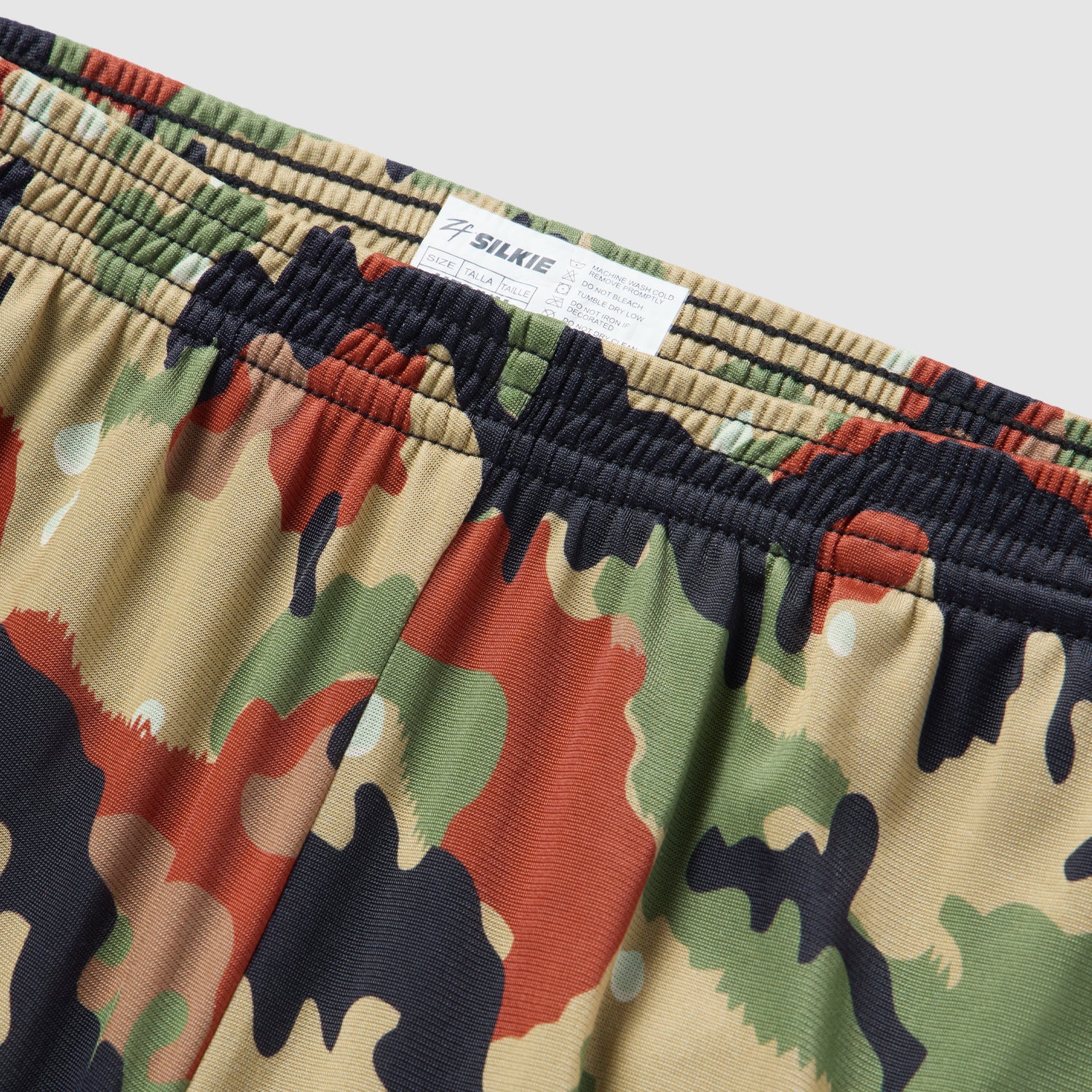 Close-up view of camo patterned shorts with a multicolor design including green, tan, black, and rust shades, featuring an elastic waistband and a tag showing the brand ZF Silkies - Camos.