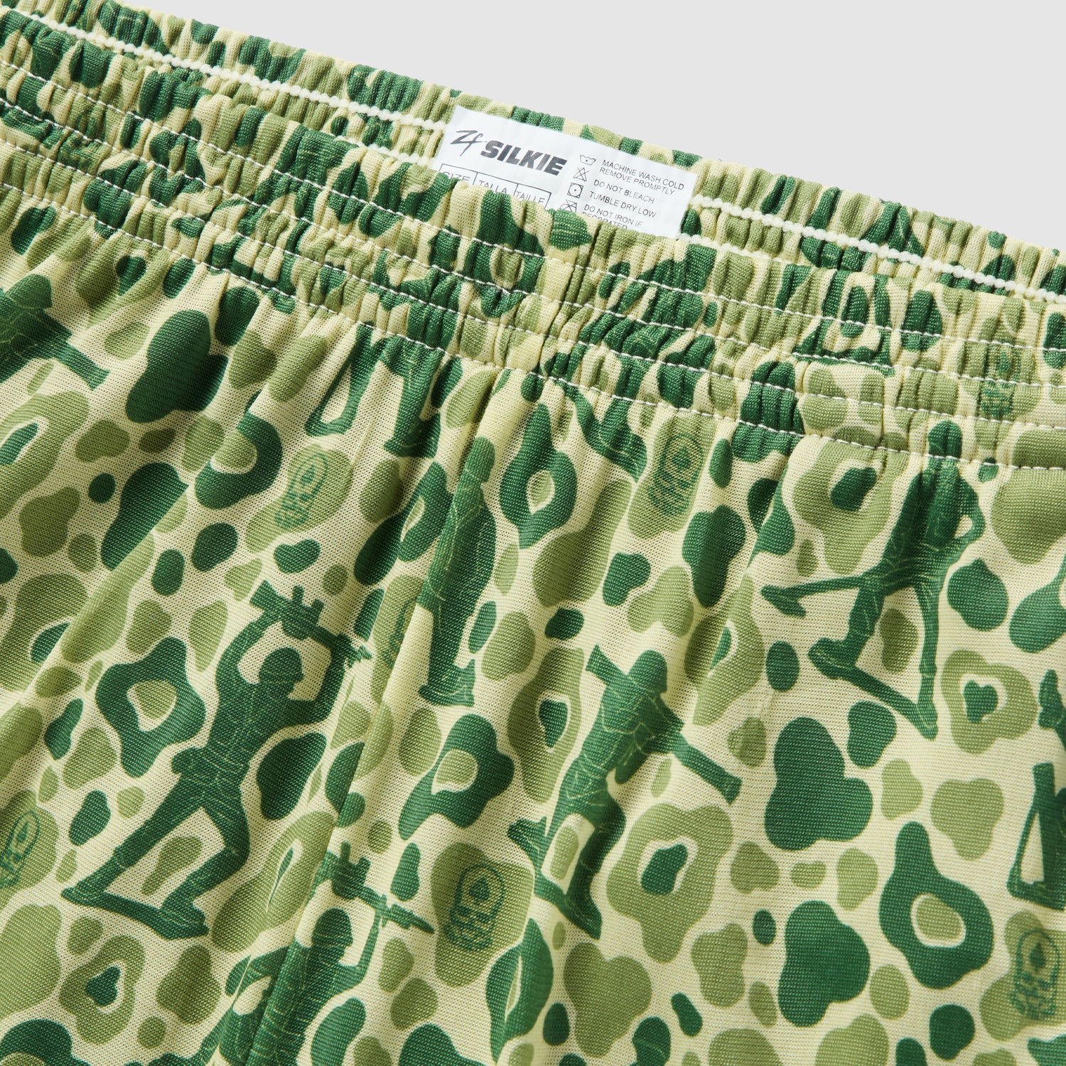 Close-up view of green and beige patterned fabric featuring green army men silhouettes with an elastic waistband, part of the Army Men Silkies shorts.